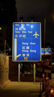 A blue highway sign is prominently displayed at night, directing travelers to various locations such as Yau Ma Tei, Tsim Sha Tsui, Hung Hom, and Hong Kong. The sign includes an airplane symbol, indicating directions to an airport. It is illuminated by nearby streetlights and is positioned on a sidewalk beside a fenced construction area.