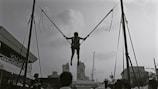 A person is suspended in mid-air, connected to two ropes and harnessed on a bungee trampoline setup. The silhouette of distant buildings and cranes is visible in the background. The event seems to be taking place outdoors, with tents and several people on the ground looking at the jumper.