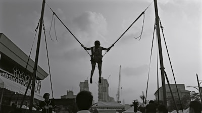A person is suspended in mid-air, connected to two ropes and harnessed on a bungee trampoline setup. The silhouette of distant buildings and cranes is visible in the background. The event seems to be taking place outdoors, with tents and several people on the ground looking at the jumper.