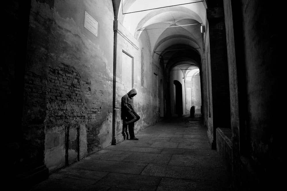 Foto en escala de grises de un hombre con sudadera con capucha apoyado en la pared del callejón