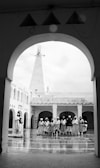 A group of people, dressed in similar traditional attire, stand in a semi-circle within a courtyard surrounded by arched architecture. The floor appears to be reflective, suggesting it might be wet. Above the courtyard, a tall, pointed structure is visible, possibly a minaret.