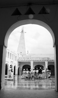 A group of people, dressed in similar traditional attire, stand in a semi-circle within a courtyard surrounded by arched architecture. The floor appears to be reflective, suggesting it might be wet. Above the courtyard, a tall, pointed structure is visible, possibly a minaret.