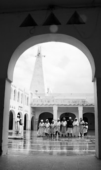 A group of people, dressed in similar traditional attire, stand in a semi-circle within a courtyard surrounded by arched architecture. The floor appears to be reflective, suggesting it might be wet. Above the courtyard, a tall, pointed structure is visible, possibly a minaret.