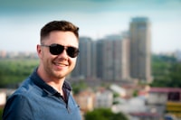 Man with glasses and a blue polo shirt standing in front of city skyline smiling