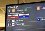 The image displays an airport check-in board featuring several airline logos and their respective check-in details. The board is predominantly black with white text and colored airline logos, showing Lufthansa prominently along with other associated airlines like SWISS, SAS, and Air Malta.