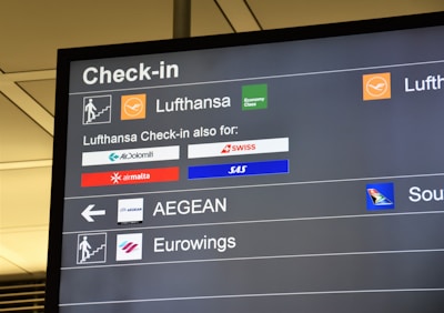 The image displays an airport check-in board featuring several airline logos and their respective check-in details. The board is predominantly black with white text and colored airline logos, showing Lufthansa prominently along with other associated airlines like SWISS, SAS, and Air Malta.