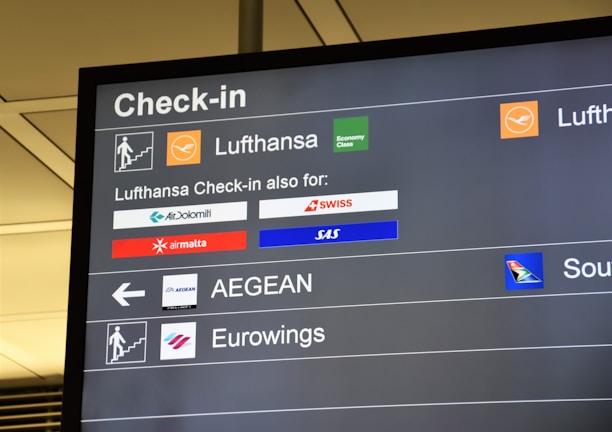 The image displays an airport check-in board featuring several airline logos and their respective check-in details. The board is predominantly black with white text and colored airline logos, showing Lufthansa prominently along with other associated airlines like SWISS, SAS, and Air Malta.