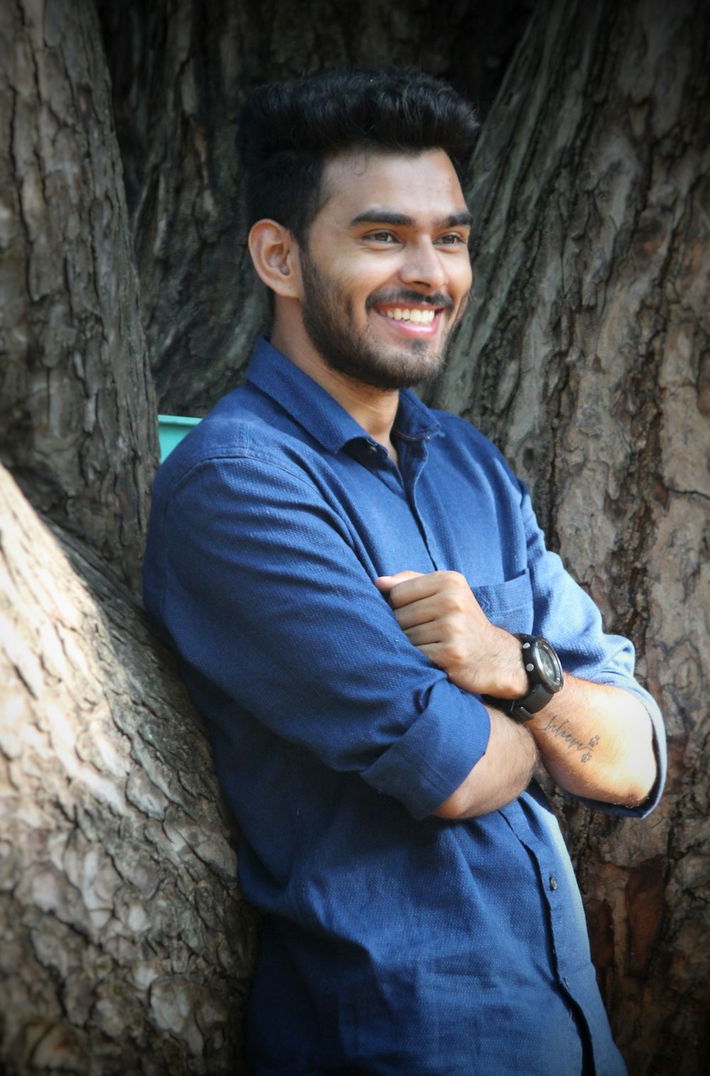 man in blue dress shirt standing and leaning on tree and smiling