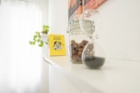 On a white shelf, there is a yellow book titled 'Nyugat Zombik', a glass jar filled with nuts, and an hourglass with black sand. A green leafy plant is also placed on the shelf, and a part of a wall painting with warm tones can be seen.