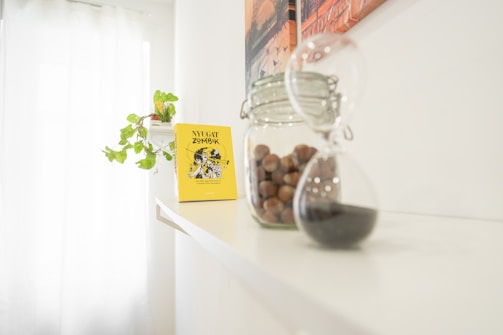 On a white shelf, there is a yellow book titled 'Nyugat Zombik', a glass jar filled with nuts, and an hourglass with black sand. A green leafy plant is also placed on the shelf, and a part of a wall painting with warm tones can be seen.