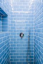 A shower area with glossy blue subway tiles covering the walls. A modern chrome showerhead is attached to a metallic pipe on the right wall, and a sleek, round temperature control is centered on the wall. The tiles are neatly aligned, creating a clean and contemporary appearance.