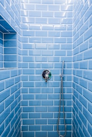 A shower area with glossy blue subway tiles covering the walls. A modern chrome showerhead is attached to a metallic pipe on the right wall, and a sleek, round temperature control is centered on the wall. The tiles are neatly aligned, creating a clean and contemporary appearance.