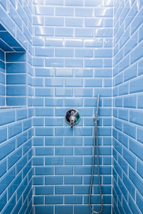 A shower area with glossy blue subway tiles covering the walls. A modern chrome showerhead is attached to a metallic pipe on the right wall, and a sleek, round temperature control is centered on the wall. The tiles are neatly aligned, creating a clean and contemporary appearance.