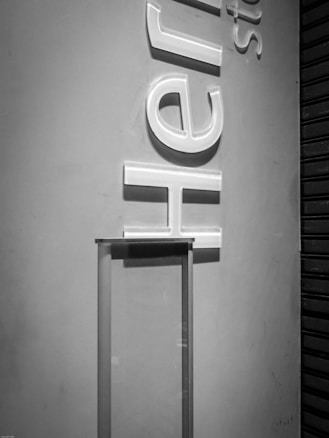 A grayscale image features a wall with large, three-dimensional letters that appear to be backlit. A partial metal stand or structure is visible in the lower center, with a vertical design that contrasts against the flat wall. The space has an industrial feel, possibly a storefront or display area.
