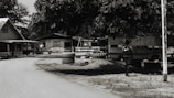 A dirt road curves gently through a small village setting, flanked by simple wooden houses with corrugated metal roofs. A large tree provides shade over an area where tires are stacked. A person with a backpack walks in the direction of a parked bus, partially visible behind the foliage.