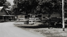 A dirt road curves gently through a small village setting, flanked by simple wooden houses with corrugated metal roofs. A large tree provides shade over an area where tires are stacked. A person with a backpack walks in the direction of a parked bus, partially visible behind the foliage.