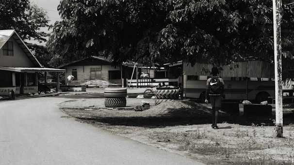 A dirt road curves gently through a small village setting, flanked by simple wooden houses with corrugated metal roofs. A large tree provides shade over an area where tires are stacked. A person with a backpack walks in the direction of a parked bus, partially visible behind the foliage.