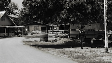 A dirt road curves gently through a small village setting, flanked by simple wooden houses with corrugated metal roofs. A large tree provides shade over an area where tires are stacked. A person with a backpack walks in the direction of a parked bus, partially visible behind the foliage.