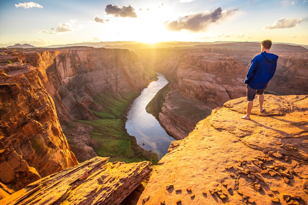 man in blue jacket standing on cliff and facing river in between rock mountain during daytime