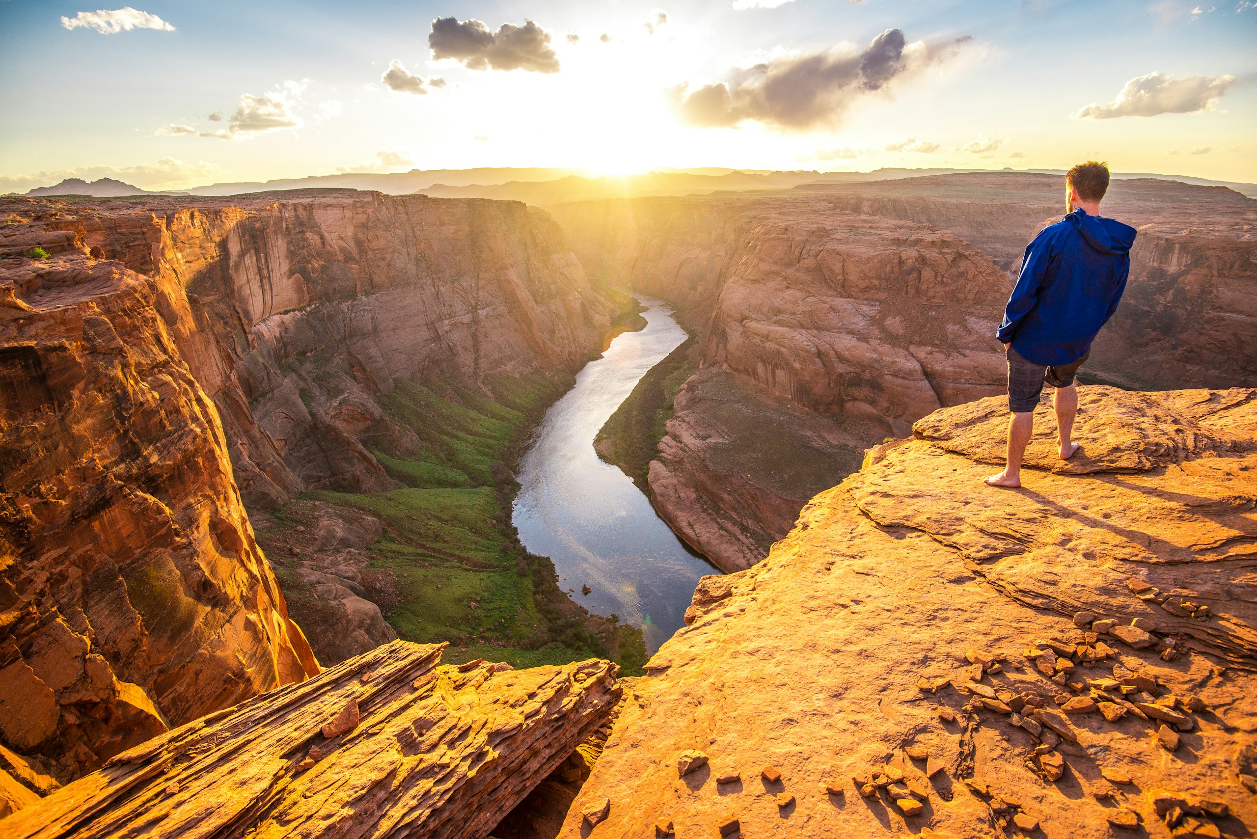 man in blue jacket standing on cliff and facing river in between rock mountain during daytime