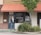 A person stands in front of a brick storefront with a large window displaying the words 'MealPro, For the Athlete in You.' A tree and some shrubs are visible on the right-hand side, and a sign prohibiting smoking is on the left. The awning above the storefront is a muted red color, and the door and window frames are dark.