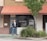 A person stands in front of a brick storefront with a large window displaying the words 'MealPro, For the Athlete in You.' A tree and some shrubs are visible on the right-hand side, and a sign prohibiting smoking is on the left. The awning above the storefront is a muted red color, and the door and window frames are dark.