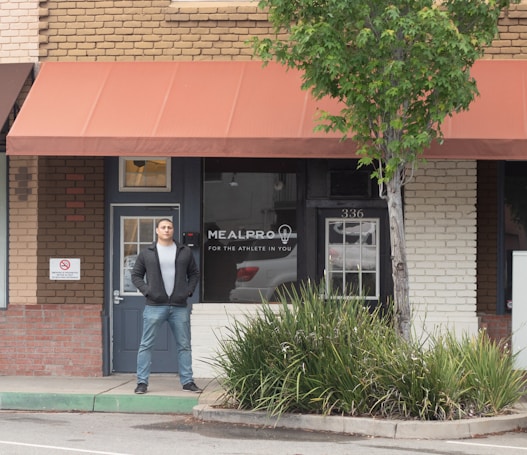 A person stands in front of a brick storefront with a large window displaying the words 'MealPro, For the Athlete in You.' A tree and some shrubs are visible on the right-hand side, and a sign prohibiting smoking is on the left. The awning above the storefront is a muted red color, and the door and window frames are dark.