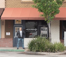 A person stands in front of a brick storefront with a large window displaying the words 'MealPro, For the Athlete in You.' A tree and some shrubs are visible on the right-hand side, and a sign prohibiting smoking is on the left. The awning above the storefront is a muted red color, and the door and window frames are dark.