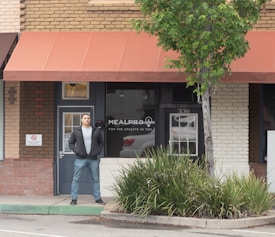 A person stands in front of a brick storefront with a large window displaying the words 'MealPro, For the Athlete in You.' A tree and some shrubs are visible on the right-hand side, and a sign prohibiting smoking is on the left. The awning above the storefront is a muted red color, and the door and window frames are dark.