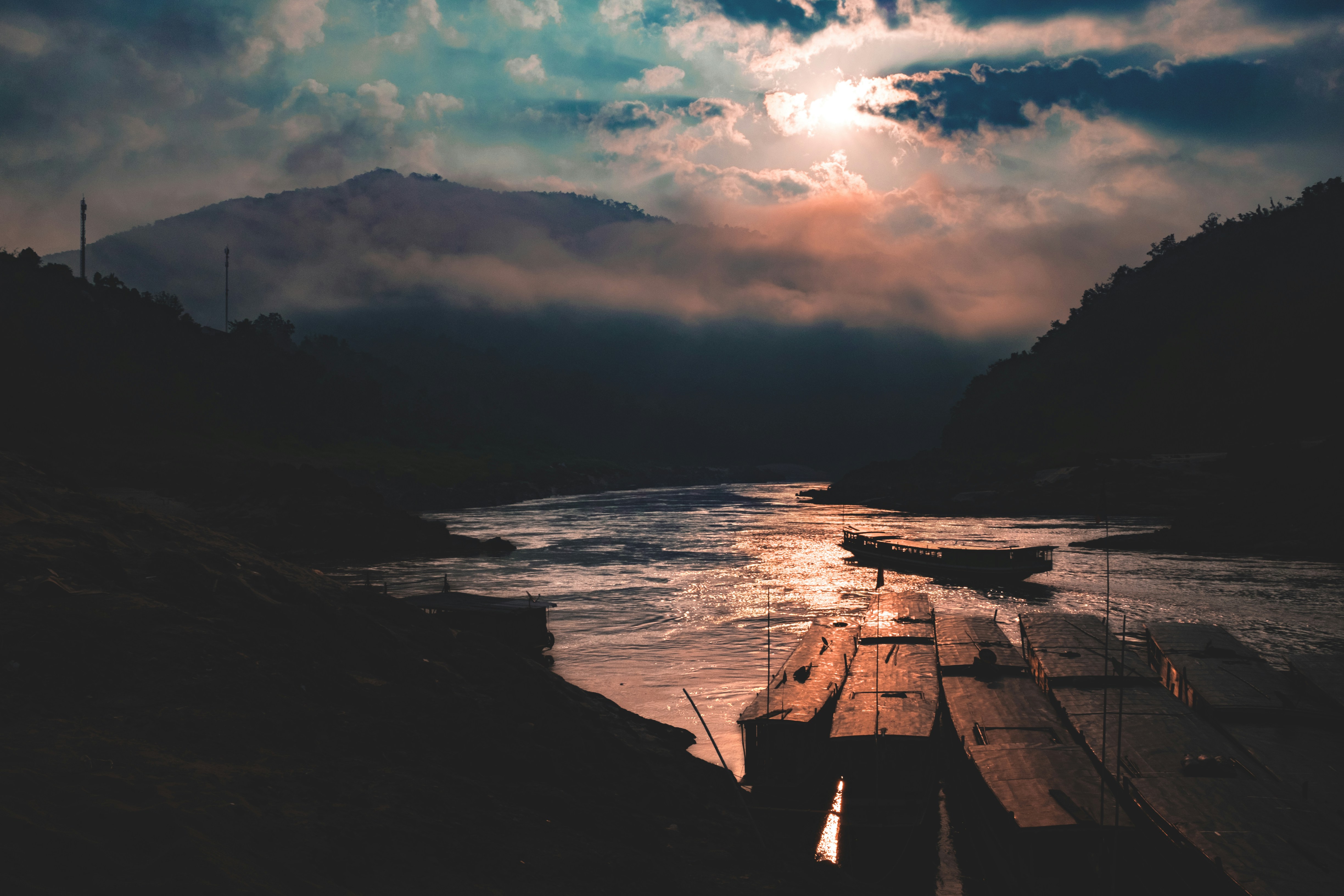 a boat sitting on top of a river under a cloudy sky, Sunset over the longboats in Luaong Probang, Laos.