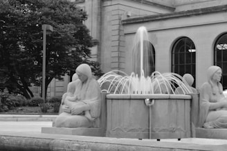 A grayscale photograph of a fountain surrounded by statues of seated figures. The figures appear to be mothers holding children, carved in a classical style. The fountain is in the foreground with water elegantly arching over its edges. Behind the statues is a building with tall windows and a large tree providing some greenery.