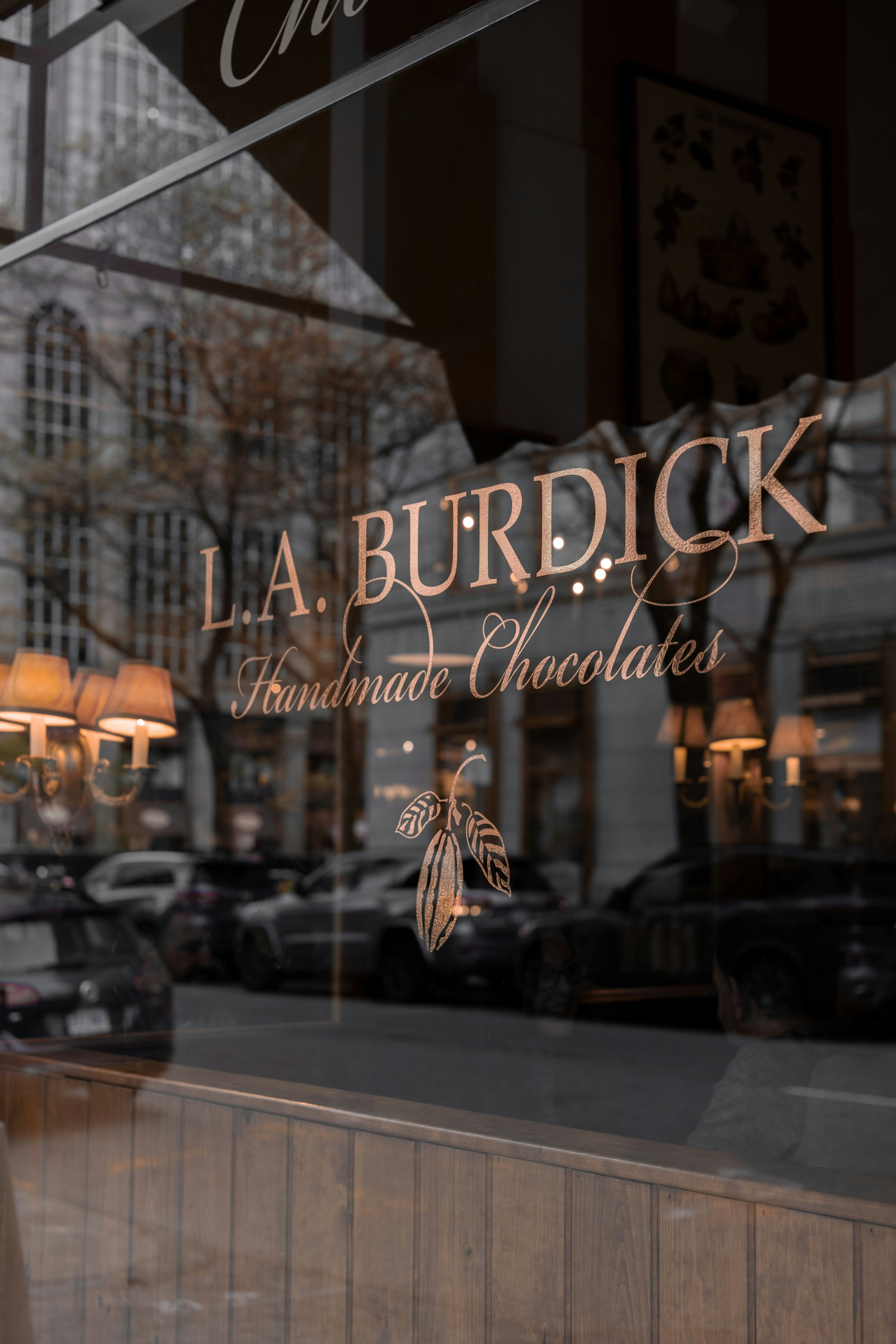 Storefront window features the L.A. Burdick logo reflected in glass. Warm interior lighting and street reflections create a layered urban scene.