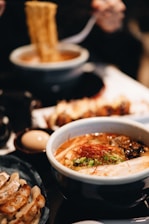 A steaming bowl of ramen is accompanied by a plate of gyoza dumplings and a soft-boiled egg. The ramen is topped with sliced green onions, red chili threads, and a slice of pork. The rich broth and noodles are being lifted with chopsticks.