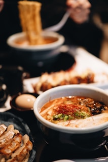 A steaming bowl of ramen is accompanied by a plate of gyoza dumplings and a soft-boiled egg. The ramen is topped with sliced green onions, red chili threads, and a slice of pork. The rich broth and noodles are being lifted with chopsticks.