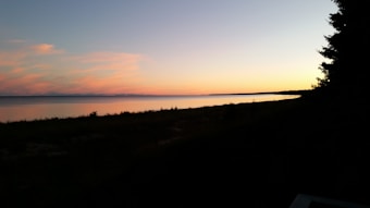 A serene landscape depicting a calm body of water reflecting the colors of a sunset. The sky is painted with gradient hues ranging from orange to blue, and the silhouette of pine trees is visible on the right side. The horizon is smooth and unobstructed.