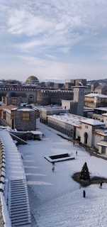 A snow-covered historical cityscape featuring a mix of ancient and medieval architecture with stone walls and towers. Several people walk along the snowy paths, and a tall Christmas tree stands decorated in an open square. The sky is partly cloudy, adding to the wintry atmosphere.