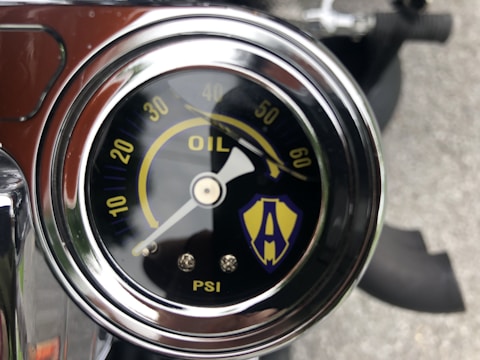 A close-up view of an oil pressure gauge with a metallic casing. The gauge displays measurements in PSI, with numbers ranging from 0 to 60. An arrow points around the 0-10 range. There is a logo or symbol featuring an 'A' on the gauge face, with a combination of yellow and blue colors.