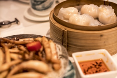 A bamboo steamer filled with white dumplings sits on a table alongside a dish of mixed stir-fried vegetables and an orange dipping sauce in a square bowl. The table setting includes a teacup and a pair of chopsticks.