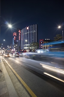 The image captures a vibrant city street at night with a long exposure effect. Tall buildings are adorned with bright neon signs and vertical lights, creating an urban skyline. A street bustling with traffic is seen on the left, and the motion blur of car lights suggests speed and activity. The scene is illuminated by streetlights casting a soft glow over the area.