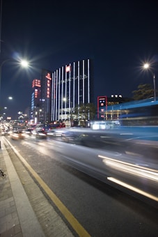 The image captures a vibrant city street at night with a long exposure effect. Tall buildings are adorned with bright neon signs and vertical lights, creating an urban skyline. A street bustling with traffic is seen on the left, and the motion blur of car lights suggests speed and activity. The scene is illuminated by streetlights casting a soft glow over the area.