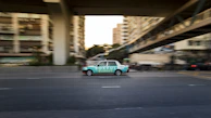A moving taxi with advertising on its side travels along a multi-lane city road, surrounded by blurred buildings and overpasses, suggesting urban motion and speed.