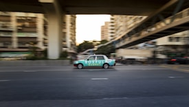 A moving taxi with advertising on its side travels along a multi-lane city road, surrounded by blurred buildings and overpasses, suggesting urban motion and speed.