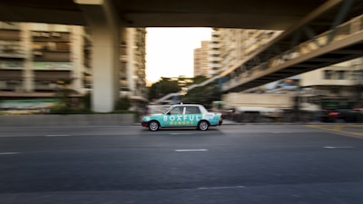 A moving taxi with advertising on its side travels along a multi-lane city road, surrounded by blurred buildings and overpasses, suggesting urban motion and speed.