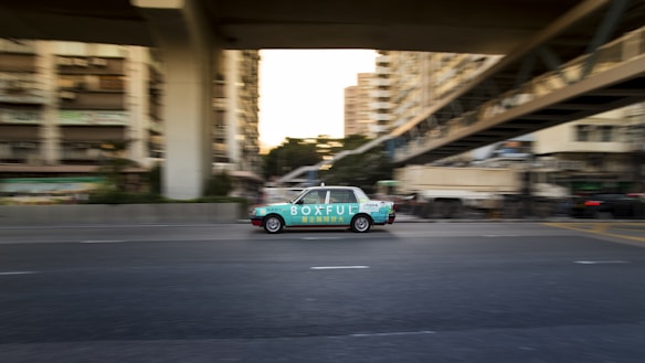 A moving taxi with advertising on its side travels along a multi-lane city road, surrounded by blurred buildings and overpasses, suggesting urban motion and speed.