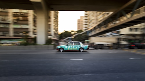 A moving taxi with advertising on its side travels along a multi-lane city road, surrounded by blurred buildings and overpasses, suggesting urban motion and speed.