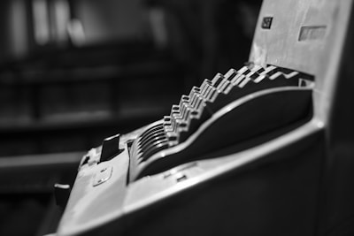 A close-up view of an old mechanical cash register with prominent levers or keys arranged in a diagonal pattern. The image focuses on the mechanical components, illustrating a tactile, vintage aesthetic. The photo is captured in black and white, emphasizing the shadows and fine details of the machine.