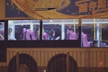 A bus with large windows revealing passengers seated inside. The interior features pink seats and yellow handrails, while the exterior is adorned with colorful advertising and Chinese text. It is nighttime, adding to the dimly lit ambiance visible through the windows.