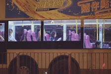 A bus with large windows revealing passengers seated inside. The interior features pink seats and yellow handrails, while the exterior is adorned with colorful advertising and Chinese text. It is nighttime, adding to the dimly lit ambiance visible through the windows.