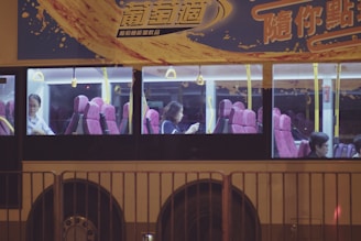 A bus with large windows revealing passengers seated inside. The interior features pink seats and yellow handrails, while the exterior is adorned with colorful advertising and Chinese text. It is nighttime, adding to the dimly lit ambiance visible through the windows.