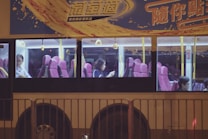 A bus with large windows revealing passengers seated inside. The interior features pink seats and yellow handrails, while the exterior is adorned with colorful advertising and Chinese text. It is nighttime, adding to the dimly lit ambiance visible through the windows.